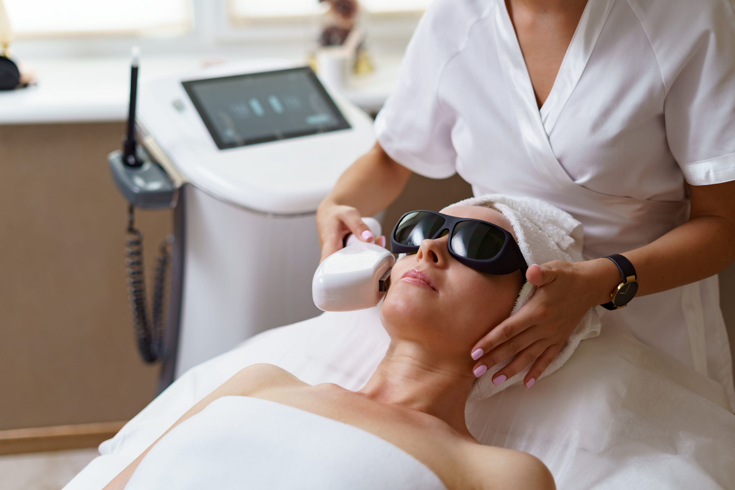 View of doctor cosmetologist doing anti aging procedure in cosmetology office. Satisfied woman in disposable hat lying on couch and relaxing. Working with Apparatus.